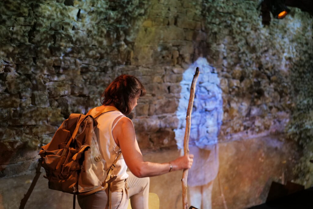 Histoire d'une marche en plein ciel — Marie-Aude Christianne au plateau. Elle tient un bâton de marche et est agenouillée devant une silhouette projetée.