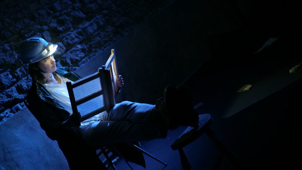 Histoire d'une marche en plein ciel — Marie-Aude Christianne au plateau. Elle porte un casque de mineur et tient une brochure. Elle est assise, les pieds reposant sur une chaise.